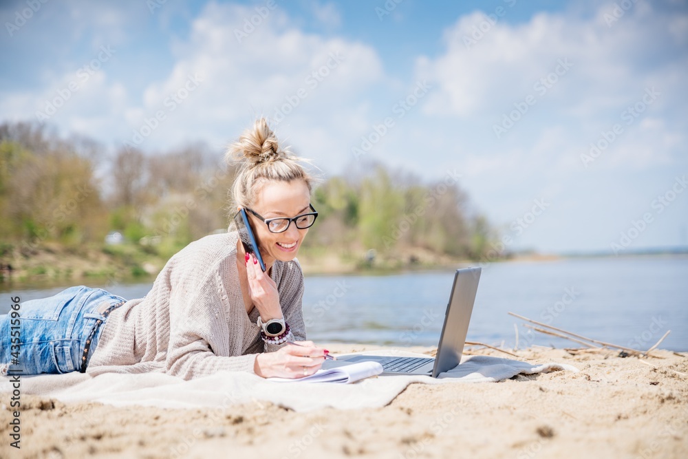 Beauty smiling woman using laptop computer on a beach. Girl freelancer working by a lake. Freelance work, travel, vacations, stay connected.