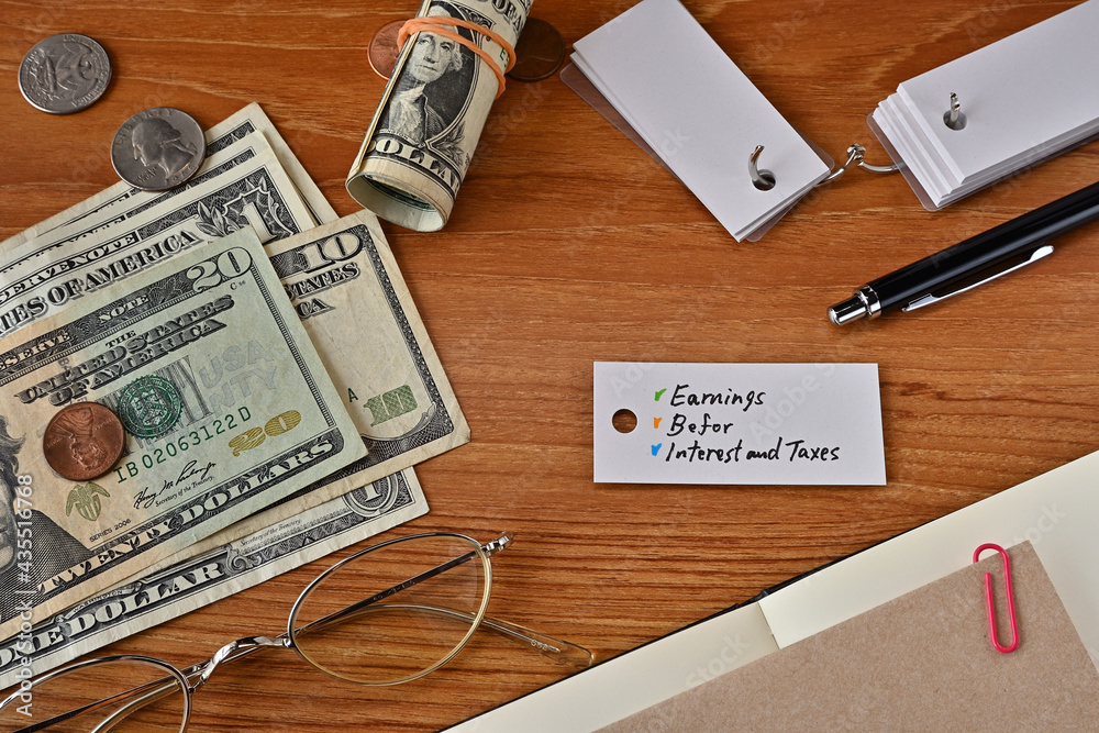 There is a word book with the word Earnings Before Interest and Taxes which is a financial term written on it. It is on a desk with bills, glasses and notebook. There is a word book with the word Earnings Before Interest and Taxes which is a financial term written on it. It is on a desk with bills, glasses and notebook.