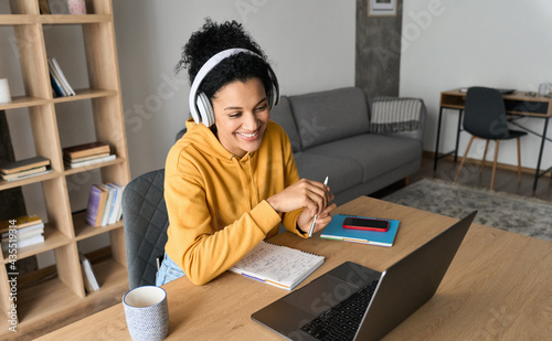 Young happy smiling African American adult student wearing headphones having virtual education class meeting online call e learning webinar on laptop at home office writing notes.