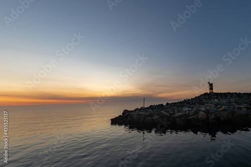 An evening sky at the horizon level with orange, red and yellow at sunset. There's land or an island in the foreground surrounded by calm smooth ocean water. The land has a small light beacon.