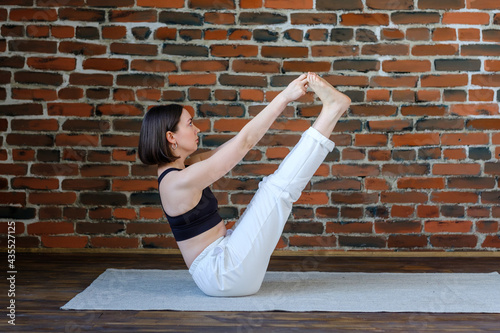 Sporty beautiful young woman in white sportswear doing exercises for legs and abs, sitting in Ubhaya Padangusthasana asana, Both Big Toe pose, side view, brickwork wall in yoga studio. photo