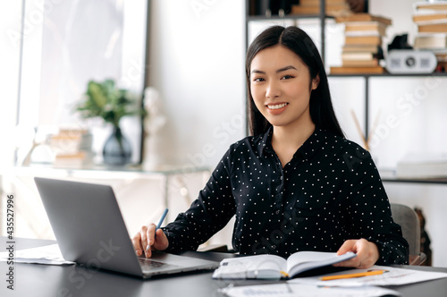 Portrait of a pretty asian young woman. Successful confident japanese female office worker, wearing formal shirt, sitting at her workplace with laptop, looking at the camera, smiling friendly