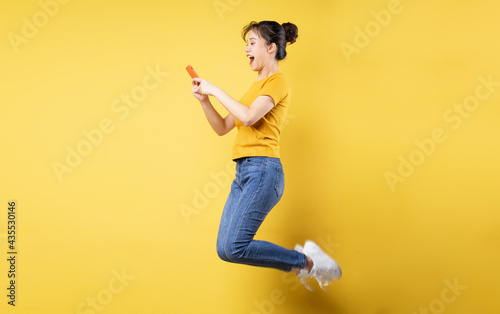 Full body profile photo of young asian girl jumping high holding a phone writing a new social media post, isolated on blue background © 1112000