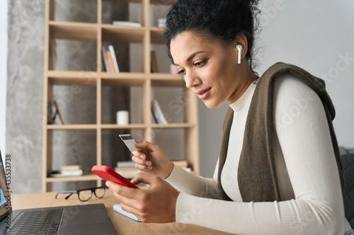 Young adult African American female consumer holding credit card and smartphone sitting at desk at home doing online banking transaction. E commerce virtual shopping, mobile banking concept. photo
