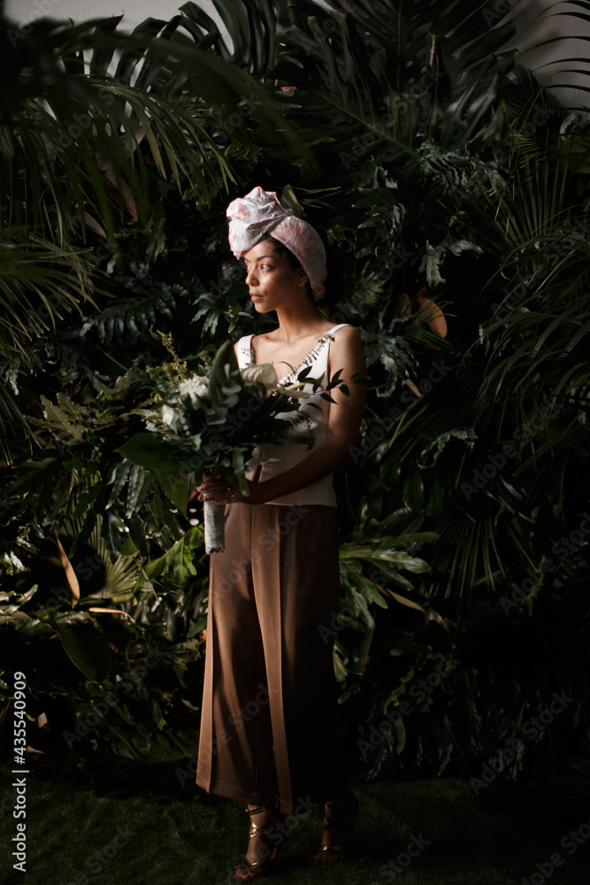 Charming calm curly hair, dark - skinned woman in white dress posing in a tropical decoration, holding a bouquet of colorful flowers in her hands Charming calm curly hair, dark - skinned woman in white dress posing in a tropical decoration, holding a bouquet of colorful flowers in her hands