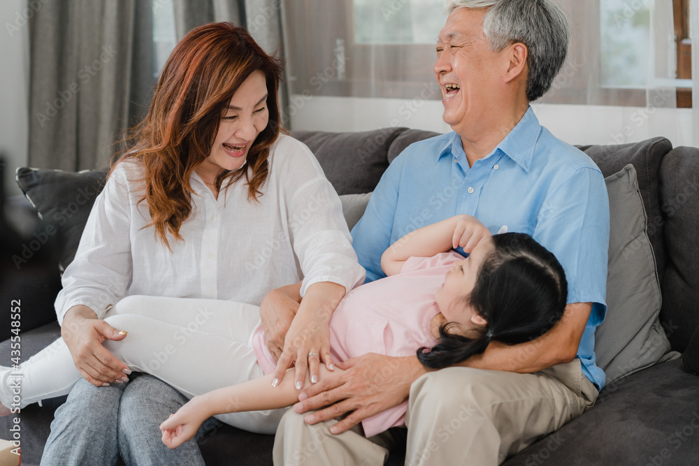 Fototapeta premium Asian grandparents talking with granddaughter at home. Senior Chinese, old generation, grandfather and grandmother using family time relax with young girl kid lying on sofa in living room concept.