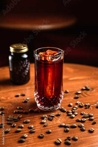 A cocktail in a collins highball glass with an ice spear, coffee beans around, a glass jar with words COFFEE BEANS on it, on a wooden table, at the bar.