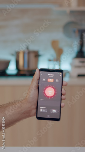 Male holding mobile with lighting control app sitting in kitchen of house with automation light system, turning on bulb. Smartphone for house automation system, high tech application for smart home photo