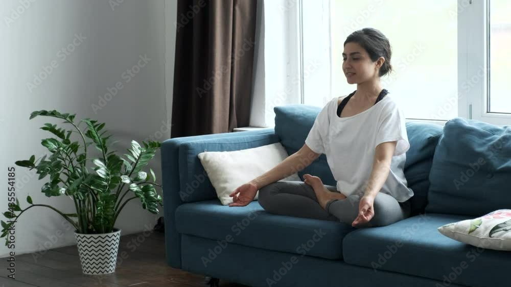 A Young Indian Woman Is Doing Yoga At Home, Sitting on a Sofa in Lotus Position in a Bright Room with Beautiful Furniture Around, The Girl Is Dressed in a White T-Shirt and Leggings A Young Indian Woman Is Doing Yoga At Home, Sitting on a Sofa in Lotus Position in a Bright Room with Beautiful Furniture Around, The Girl Is Dressed in a White T-Shirt and Leggings