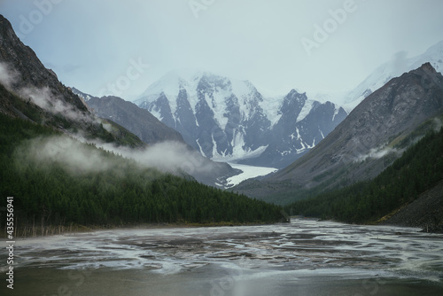 Atmospheric alpine landscape with mountain lake with streams from snowy mountains in overcast weather. Gloomy mountain scenery with green lake with rainy circles and low clouds in mountain valley.