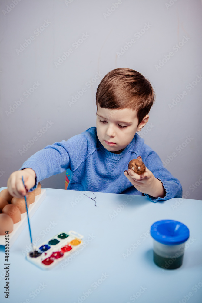 A little child paints an easter egg at the table on a blue background.Little Boy painting Easter Eggs with Paint at Home. Children's Easter creativity. A child decorate Easter eggs. A little child paints an easter egg at the table on a blue background.Little Boy painting Easter Eggs with Paint at Home. Children's Easter creativity. A child decorate Easter eggs.