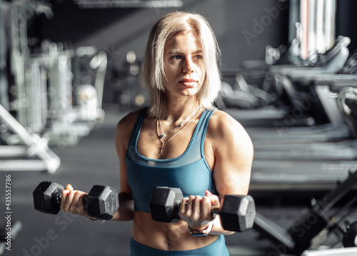Athletic woman in sportswear trains biceps with heavy dumbbells in her hands at gym. Healthy lifestyle, fitness and bodybuilding concept.