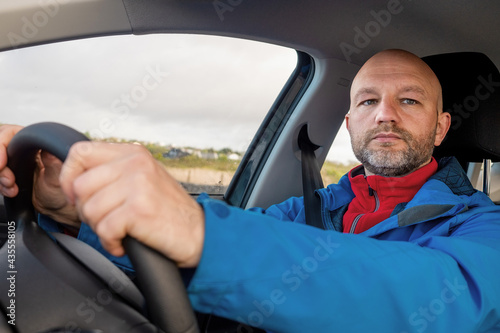 Tourist male driver in a blue jacket driving a car. The model is in his 40s, bald, grey short beard.