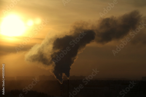 Sun below the horizon and clouds in the fiery dramatic orange sky at sunset or dawn backlit by the sun. City panorama in a foggy haze, smoke from a chimney. Place for text and design.
