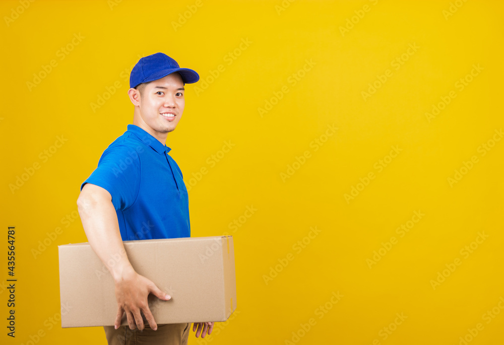 Portrait excited attractive delivery happy man logistic standing smile wearing blue t-shirt and cap uniform holding parcel box looking to camera, studio shot isolated on yellow background, side view Portrait excited attractive delivery happy man logistic standing smile wearing blue t-shirt and cap uniform holding parcel box looking to camera, studio shot isolated on yellow background, side view