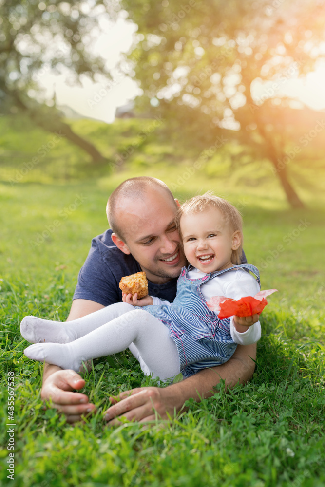 A little girl is eating a pie in her father's arms and laughing merrily. The relationship between father and daughter. Picnic in the park at sunset. A loving father. Happy family. Childhood in harmony