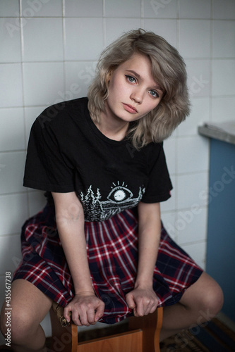 young attractive caucasian female with short hair in black t-shirt and checkered skirt sitting on a wooden stool near a wall covered with tile