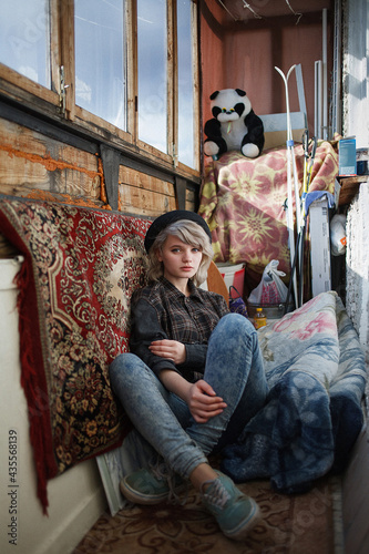 young attractive caucasian female with short hair in checkered shirt, blue jeans and black hat sitting at old vintage balcony among mess, surrounded by different junk