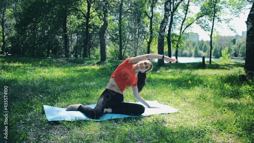 A woman in the forest does a back stretch while sitting on a mat in the park (forest) by the lake. The camera is still there. You can see the lake in the background.The general plan. Against the sun photo