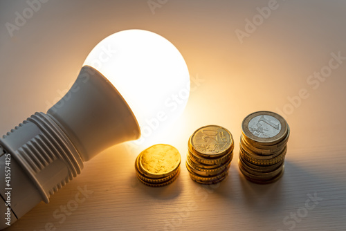 Led light bulb on, with coins beside it, on a white surface. Cost of energy, increase in energy tariffs 