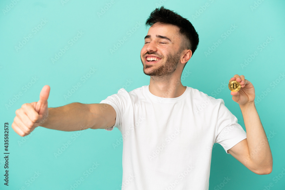 Young man holding a Bitcoin isolated on blue background giving a thumbs up gesture Young man holding a Bitcoin isolated on blue background giving a thumbs up gesture