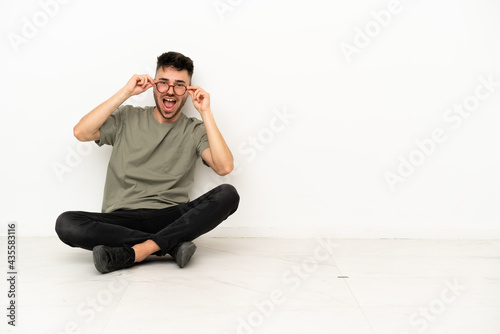 Young caucasian man sitting on the floor isolated on white background with glasses and surprised