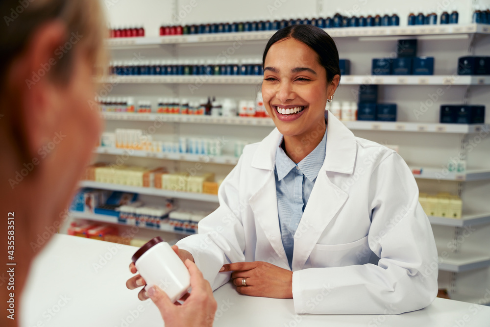 Happy female pharmacist wearing labcoat and standing behind counter giving bottle of pills to customer