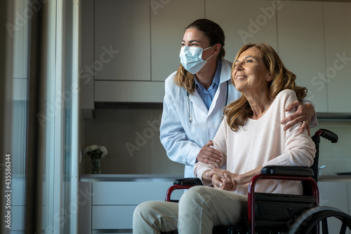 Cinematic shot of social worker with medical mask taking care of senior woman in wheelchair while looking through window at home. Concept of healthcare, protection, assistance, caregiver, covid-19. photo