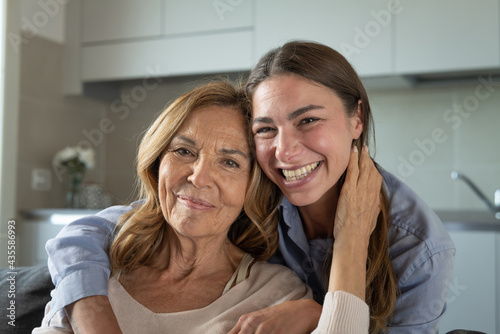 Cinematic portrait of happy grandmother and granddaughter embracing with affection and smiling in camera while sitting on sofa at home. Concept of life, grandparents, generation, love, care, family