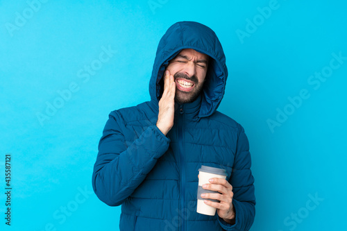 Man wearing winter jacket and holding a takeaway coffee over isolated blue background with toothache