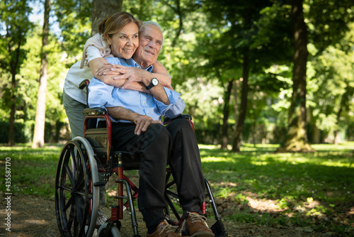 Cinematic shot of happy smiling mature wife is giving affective hug to disable husband in wheelchair as sign of forever timeless love in green park. Concept of life, family, marriage, happiness, age.