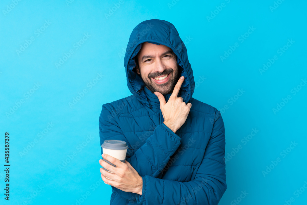 Man wearing winter jacket and holding a takeaway coffee over isolated blue background smiling