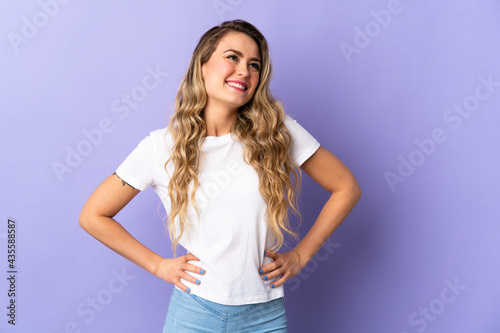 Young Brazilian woman isolated on purple background posing with arms at hip and smiling © luismolinero