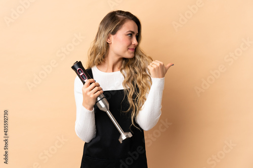 Young brazilian woman using hand blender isolated on beige background pointing to the side to present a product