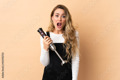 Young brazilian woman using hand blender isolated on beige background with surprise facial expression