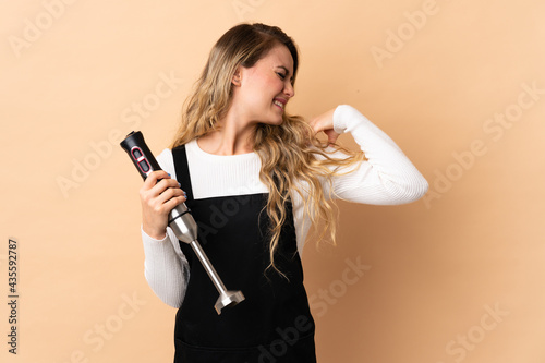 Young brazilian woman using hand blender isolated on beige background suffering from pain in shoulder for having made an effort