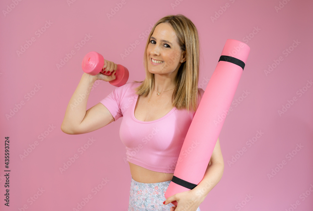 Cute athletic woman in sportswear with yoga mat and dumbbell isolated on pink background Cute athletic woman in sportswear with yoga mat and dumbbell isolated on pink background