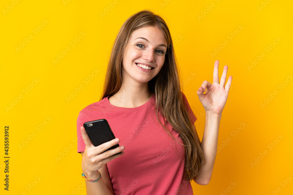 Young woman using mobile phone isolated on yellow background showing ok sign with fingers Young woman using mobile phone isolated on yellow background showing ok sign with fingers