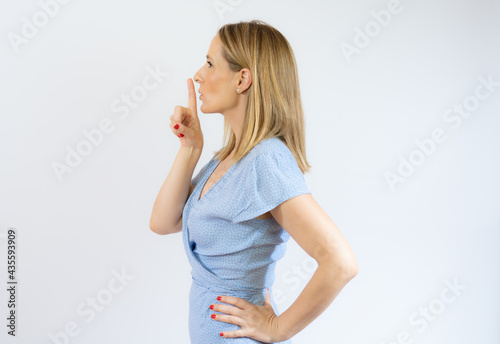 Side view young woman showing hush gesture holding index finger near lips while standing on white studio background with copy space.