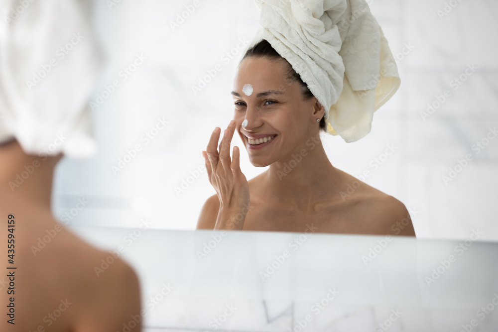 Happy millennial girl enjoying home spa, applying cream, cleaning mask on smiling face, taking care about smooth facial skin after shower, looking at reflection in mirror. Cosmetology concept