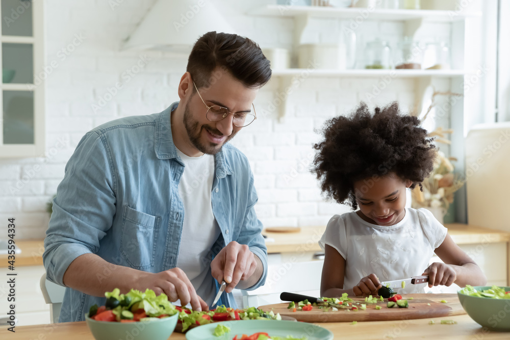 Loving young Caucasian father and small African American daughter have fun cook healthy salad together. Happy caring dad and little girl child prepare vegetarian food. Adoption, custody concept.