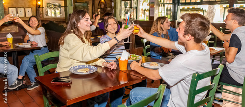 Group of millennials friends eating at fast food indoors in irish pub restaurant. Happy people partying and making a toast. Young active teenagers with burgers, pizza and drinks having fun dining.