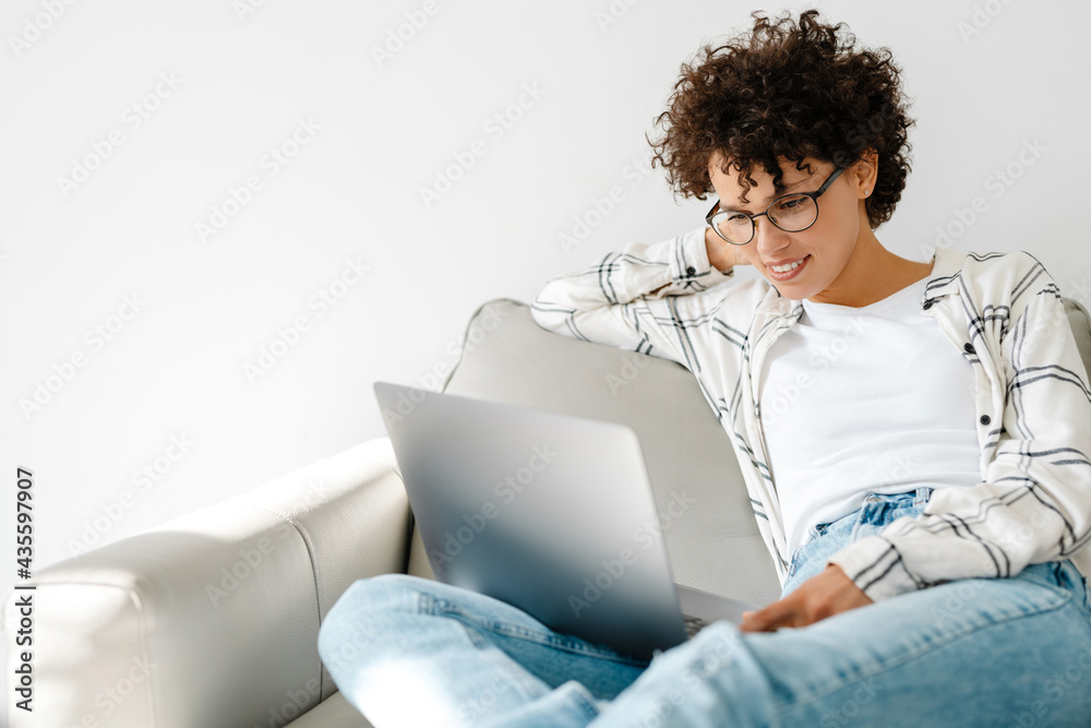 Young curly woman smiling and using laptop while sitting on couch Young curly woman smiling and using laptop while sitting on couch
