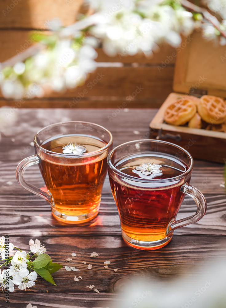 Tea party on a wooden table with branches of blossoming cherry. Black tea in glass mugs. Cherry flowers in a cup. Wooden chest with cookies in the background. Side view. Tea party on a wooden table with branches of blossoming cherry. Black tea in glass mugs. Cherry flowers in a cup. Wooden chest with cookies in the background. Side view.