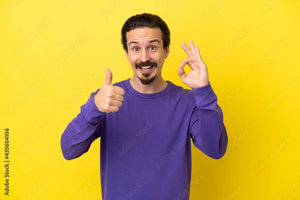 Young caucasian man isolated on yellow background showing ok sign and thumb up gesture