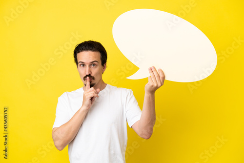 Young caucasian man isolated on yellow background holding an empty speech bubble and doing silence gesture
