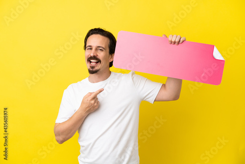 Young caucasian man isolated on yellow background holding an empty placard and pointing it