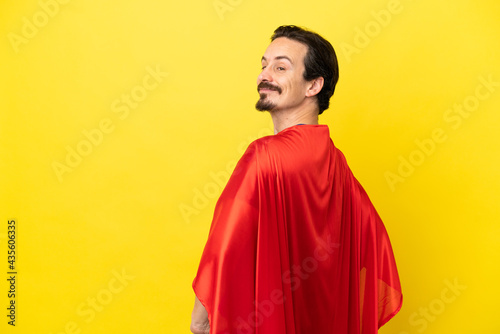 Young caucasian man isolated on yellow background in superhero costume and doingposing with arms at hip and smiling