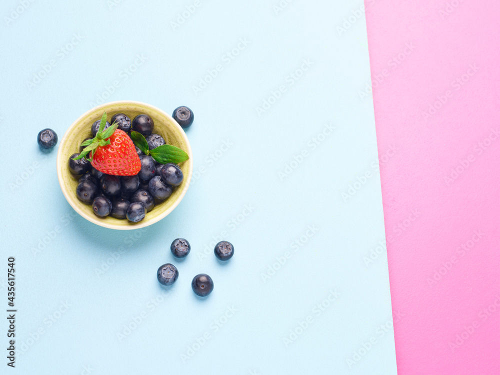 Bowl of fresh blueberries and a strawberry on a blue and pink background