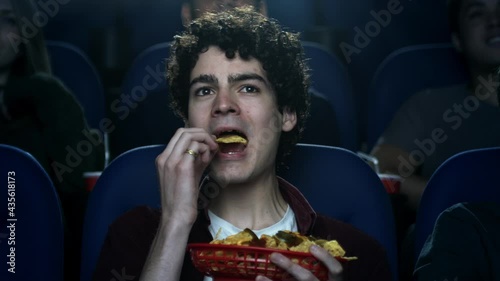 Close up of a young man sitting in a movie theatre watching a movie eating nachos.